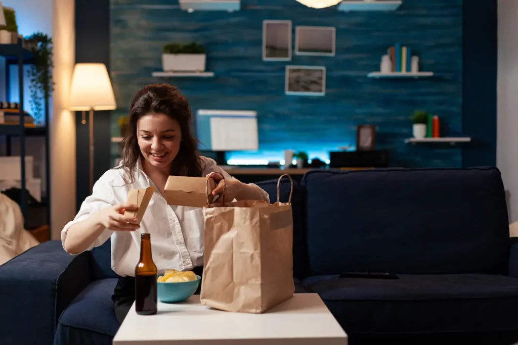 smiling woman unpacking fastfood with beer home delivered food sitting on couch